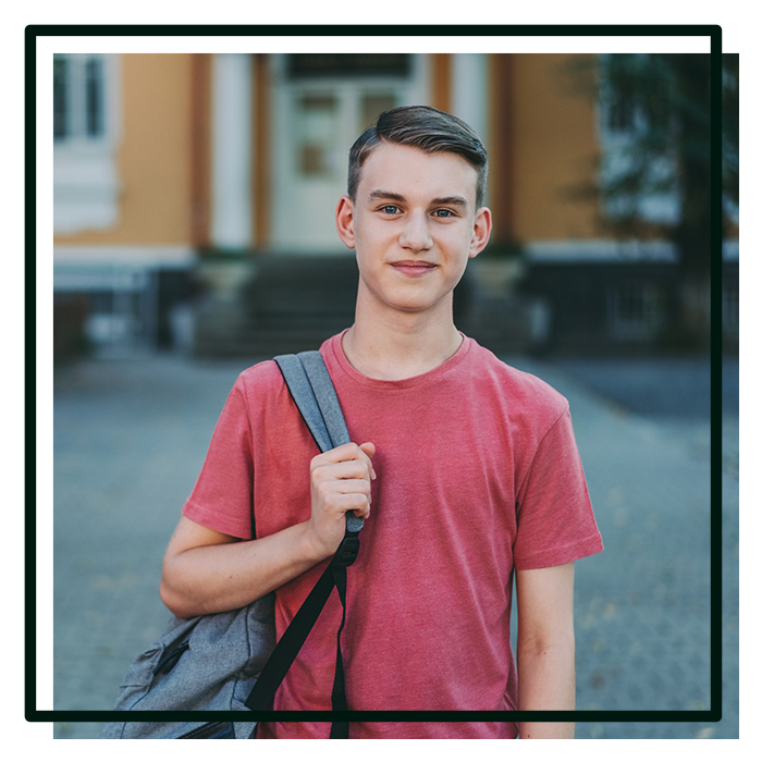 Teenage boy stands smiling holding a backpack in front of a school